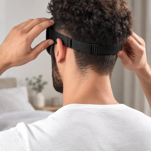 Man adjusting the elastic strap of a black 3D sleep mask from behind, natural bedroom lighting